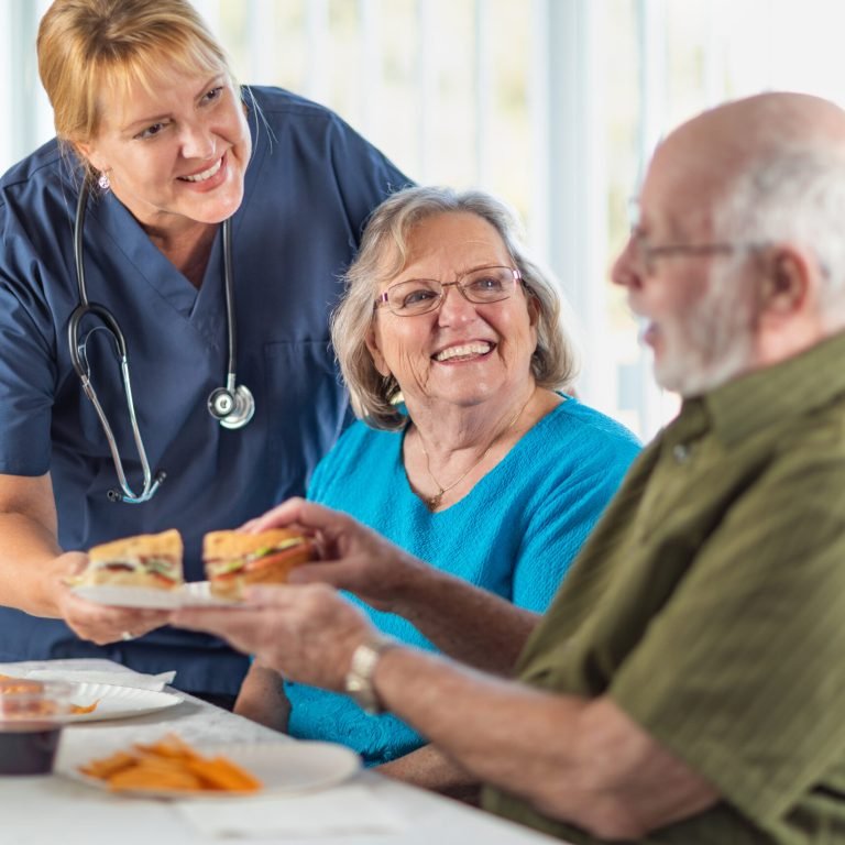 Female Doctor or Nurse Serving Senior Adult Couple Sandwiches at Table.